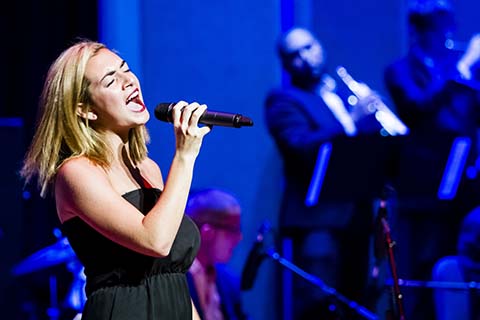 Woman vocalist in a black dress holding a microphone closes her eyes as she sings